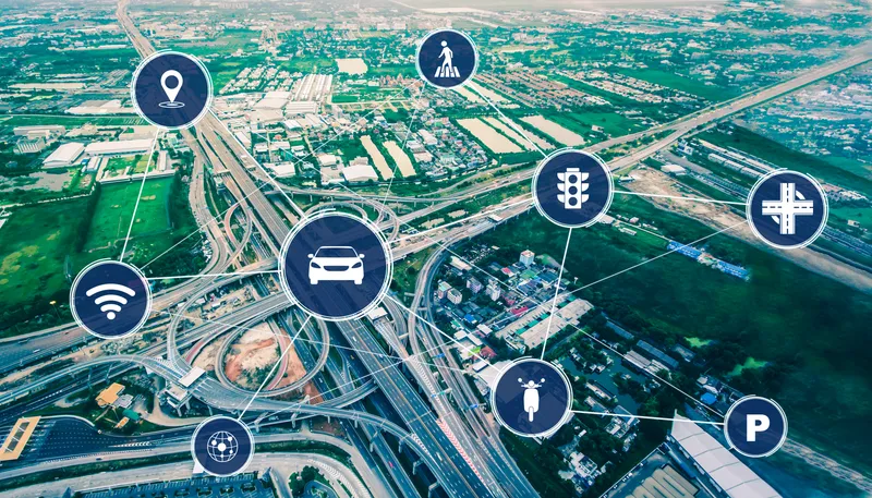 Aerial view of a complex highway interchange surrounded by urban and green areas. Several icons are overlaid on the image, connected by lines to a central car icon. The icons represent various elements related to transportation and infrastructure: a location pin, a pedestrian, traffic lights, road signs, parking, Wi-Fi signal, and a globe with network connections