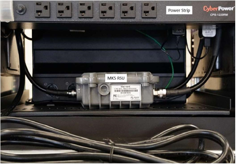 This photo is of a MK5 roadside unit (RSU) in the Connected and Automated Vehicle education (CAVe)-in-a-box infrastructure kit. The RSU is positioned below the power strip, both of which are labeled.