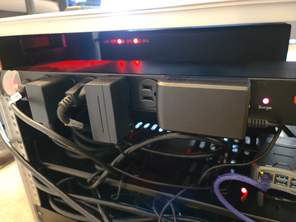 This photo shows the power strip containing eight power outlets in the CAVe-in-a-box toolkit. Various components of CAVe-in-a-box are plugged into the outlets. The surge light is on, indicating that the power strip is offering surge protection.