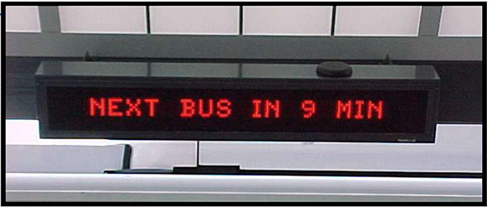 This is a photograph of a digital transportation message display board. It is a black electronic device with a red LED display. It reads: Next bus in 9 min.