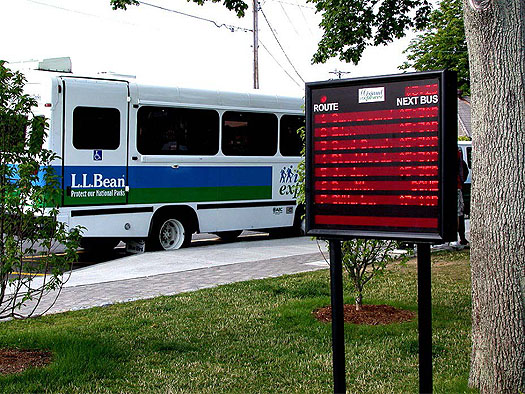 This photo represents a case study on the Island Explorer Transit ITS. In this photo, there is a large city bus parked along the side of the road. To the right of the bus is an eletext-centeronic display that show the estimated times of arrivals for various bus routes.