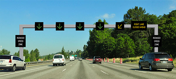 The photo shows a few cars traveling down a straight highway road and underneath overhead variable speed limit signs. There is one black speed limit sign per lane, each with a green arrow pointing down. The sign on the left side of the road reads Reduced Speed Zone. The last black speed limit sign above the lane on the far right side has a yellow arrow pointing diagonally down to the left. A final sign on the far right reads Right Lane Closed Ahead.