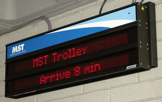 This is an electronic sign mounted on a wall at a Monterey/Salinas Transit stop. The sign is made up of two message panels. The top panel reads "MST Trolley." The bottom panel reads "Arrive 8 min."