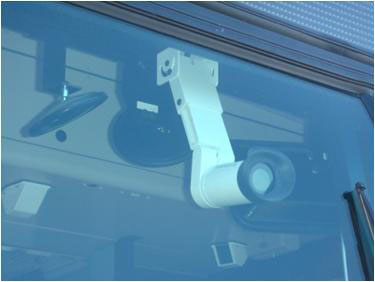 This is a photograph of an on-board surveillance camera. The camera in this photo is seen behind glass. It has a white exterior and is installed inside a vehicle via a hanging ceiling mount.