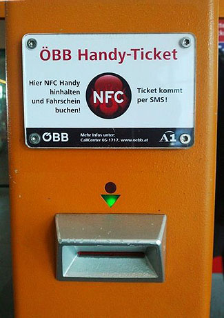 This photograph depicts a transit ticketing device from Austria. It is a large orange metal box, with a sign bolted to the front that reads OBB Handy-Ticket / Hier NFC Handy hinhalten und Farhschein buchen! / Ticket kommt per SMS! with a circular red NFC logo in the center. Below is a circular light (unlit), a triangular green light (lit), and a metal card slot at the bottom.