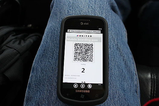 This is a photograph of a smart phone laying on a person's leg. The screen shows the United Airlines Mobile Boarding Pass application with a large QR code on the screen.