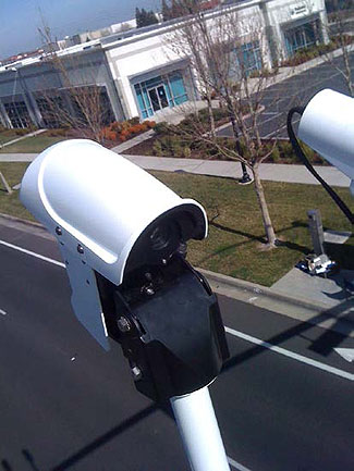 This is a photograph of a video vehicle detection unit, showing the devices high above the roadway below with buildings and a parking area in the background.