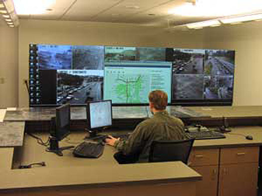 This is a photograph of a man sitting in front of a computer and a wall-sized monitor display. On the display are windows showing images from roadside surveillance cameras and maps.