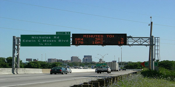 This is a photograph of cars driving under a dynamic message sign. The sign is mounted on an overhead highway beam next to a static highway road sign. The dynamic message sign is displaying distance to travel to specific destinations and estimated travel times.