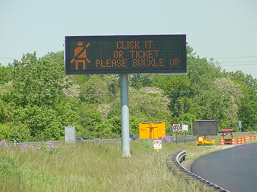 This photograph shows a full matrix dynamic message sign mounted on a post to the left of the road. The message on the sign reads: Click it or Ticket Please Buckle Up, next to an orange image of a person wearing a seatbelt.