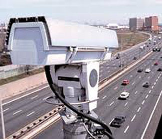This is a photograph of a white video camera mounted high above a highway. The camera is pointing down toward a highway beneath.