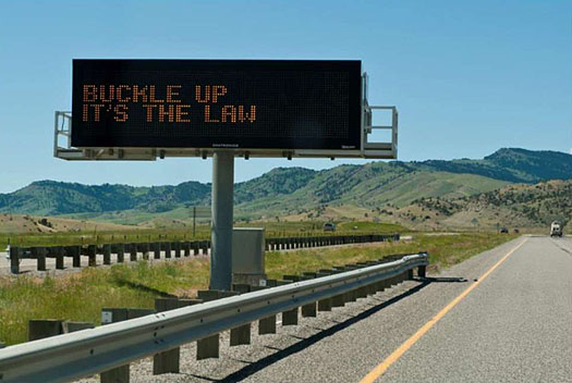 This is a photo of ITS technology along a rural road. The sign sits in the grassy divide between the two sides of the road. The sign is black, with LEDs that reads: Buckle Up It's The Law. To its right is the road with cars traveling towards the horizon.