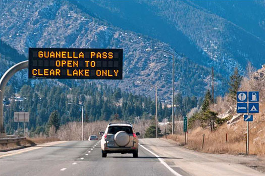 This photo illustrates how ITS technologies disseminate information for tourism and traveler. The photo shows an SUV driving towards a mountainous area. Overhead, there is an electronic message board that reads: Guanellea Pass Open to Clear Lake Only.