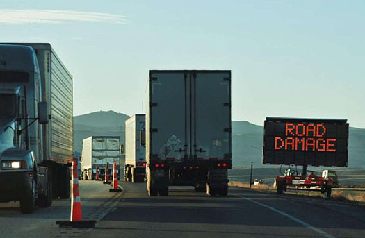 This photo illustrates how ITS technology warns travelers of infrastructure operations and maintenance. In the photo, trucks are traveling in both directions along a road that is separated by tall orange lane dividers. On the right side of the road is an electronic sign that reads: Road Damage.