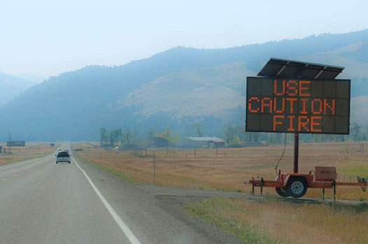 This photo illustrates how ITS technology and projects are used to warn travelers of emergencies. This image shows cars driving down a smoke-filled open road. To the right is an electronic message board that reads: Use Caution Fire.