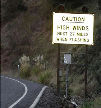 This photo represents a case study on Wind Warning Systems. In the photo, there is a white traffic sign posted with two circular lights mounted on top. The sign sits on the right side of the road and reads: Caution. High Winds next 27 miles when flashing.