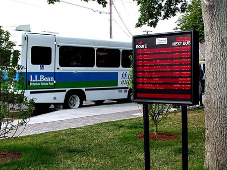 This photo represents a case study on the Island Explorer Transit ITS. In this photo, there is a large city bus parked along the side of the road. To the right of the bus is an electronic display that show the estimated times of arrivals for various bus routes.