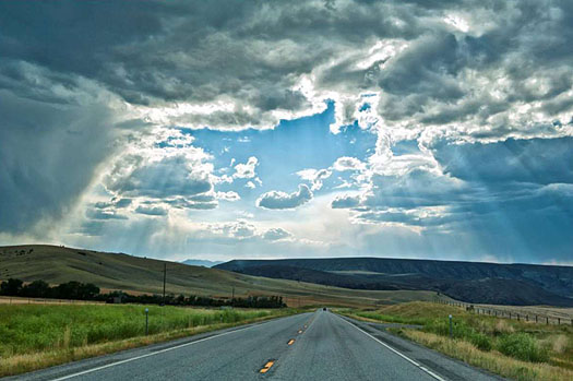 This is photo showing a rural two-lane highway heading into the distance, with a blue sky and sunshine coming through the clouds, with grassy fields on each side and hills in the background.