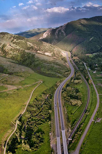 This photograph shows an aerial view of an open road in the valley of the mountains of the Western United States. There is a blue sky with white and gray clouds in the sky. Mountains are visible on the right side of the photograph as a road runs vertically in the green valley up the center and left side of the picture.