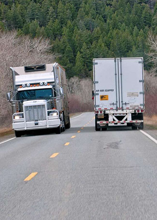 This is a photographer of two large, white 18-wheel trucks passing one another on a mountain road. On the left side of the road, the truck is facing the camera. Next to it on the left side of the road is a truck driving away from the camera.