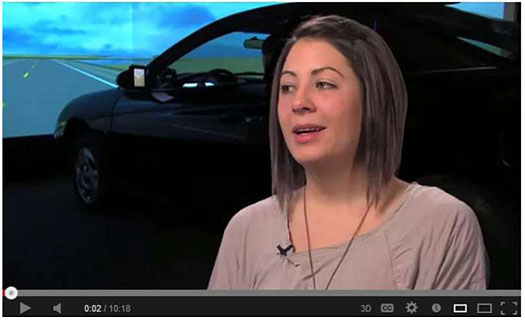 This photo is a still frame from a video from the University of Minnesota promoting careers in ITS. It shows a woman in a light brown sweater speaking in front of a backdrop showing a car in front of a large video projection of an open road.