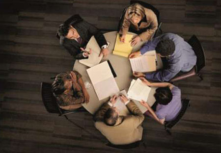 This image is an overhead photograph taken of six people having a meeting, seated around a round table with paper and notebooks.