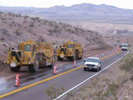 This is a photograph of two large construction trucks parked along the side of the road. The construction is coned off, and traffic is flowing towards the camera on the opposite side of the road.