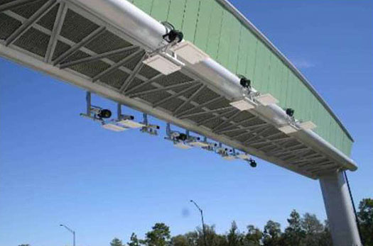 This is a photograph of overhead traffic sensors. Six sensors are mounted along a large metal beam. Three on the front edge, and three on the back edge of the beam. Cameras are arranged a few feet from each another near the sensors.