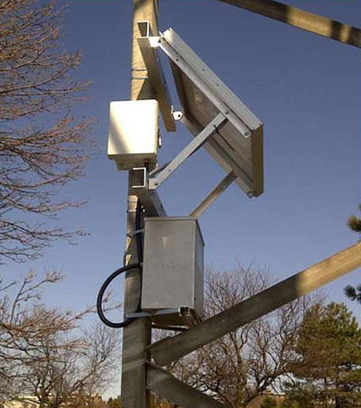This is a photograph of traffic data collection technology. Mounted on the right face of a pole is a metal box resting on an upwardly diagonal beam. Above the metal box, mounted on the front-face of the pole is a white metal box or sensor. Above the white box mounted on the right face of the pole is a metal device that is installed in a downward diagonal direction.