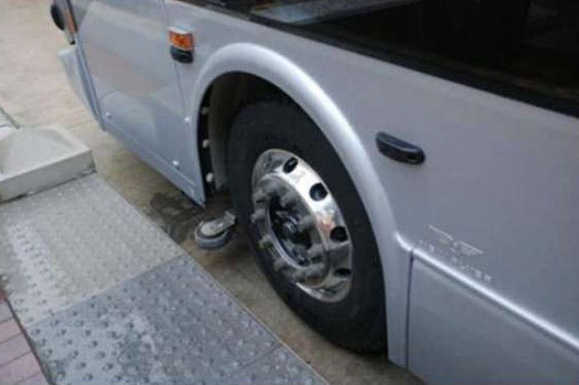 This photograph shows an example of a Vehicle Assist and Automation tool. The picture shows the front left wheel of a bus. To the left of the bus is a gray curb. Attached to the bus just in front of the wheel is a small wheel installed sideways so that the rotating portion of the little wheel extends beyond the normal width of the bus. For additional relevant information about this photo, please see the Author Notes below.