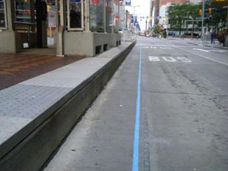 This is a photograph looking down a city street at curb level. On the left is a gray curb. To the right of the curb, there is a light blue line running along the bus lane. For additional relevant information about this photo, please see the Author Notes below.