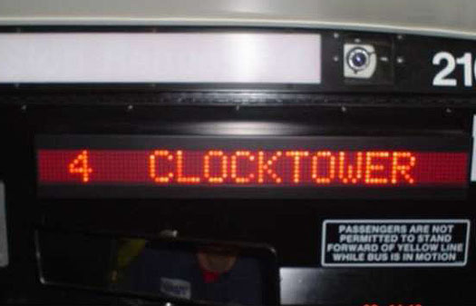 This is a photograph of a dynamic message sign inside a vehicle. The sign is installed on the ceiling of the bus, and the message appears via red LED lights. The message on the display reads: 4 Clocktower.