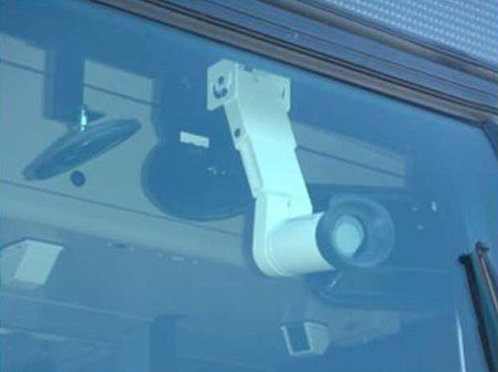 This is a photograph of an on-board surveillance camera. The camera in this photo is seen behind glass. It has a white exterior and is installed inside a vehicle via a hanging ceiling mount.
