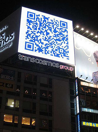 This is a photograph of a city building at night with a large digital screen at its top, next to a lighted billboard to the right, and a lighted business sign to the left. There is a blue QR code on the large digital screen and below it are the words: transcosmos group.
