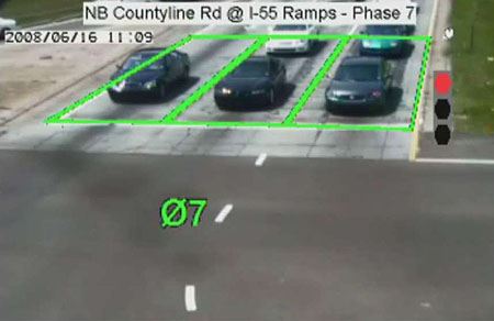 This is an example of images from a video camera sent through a Video Image Processing Unit. In this image, there are three lanes of traffic waiting at an intersection. Lane space occupied by the first vehicles in line are outlined in green. To the right is an icon of a stoplight with the red light highlighted. At the top of the image is the location of the photo as well as the light phase. Source: Econolite