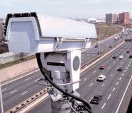 This is a photograph of a white video camera mounted high above a highway. The camera is pointing down on a highway.