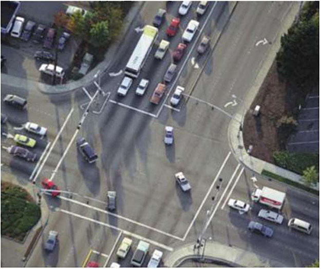 Aerial overhead photo of four-way intersection with many lanes of traffic, with numerous vehicles at a red light as some vehicles are turning.