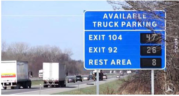 Photo example of real time truck parking information management sign along roadway in Wisconsin with some trucks on the roadway; the sign indicates the number of available truck parking spaces at Exit 104 - 47, Exit 92 - 26, and at the Rest Area - 8.