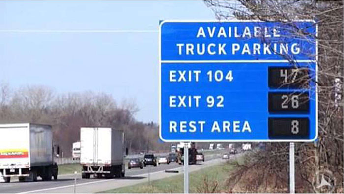 Photo example of real time truck parking information management sign along roadway in Wisconsin with some trucks on the roadway; the sign indicates the number of available truck parking spaces at Exit 104 - 47, Exit 92 - 26, and at the Rest Area - 8.