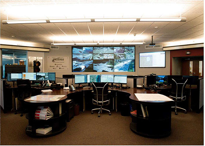 Photo showing an example county transportation management center in Lake County Illinois with a large desk and numerous computer screens and display feeds from cameras.