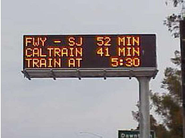 This photograph is an example of how multimodal displays are used to disseminate parking information. This is a photo of an LED message board display that is mounted on a tall metal pole. The sign reads FWY-SJ 52 min, Caltrain 41 min, Train at 5:30.