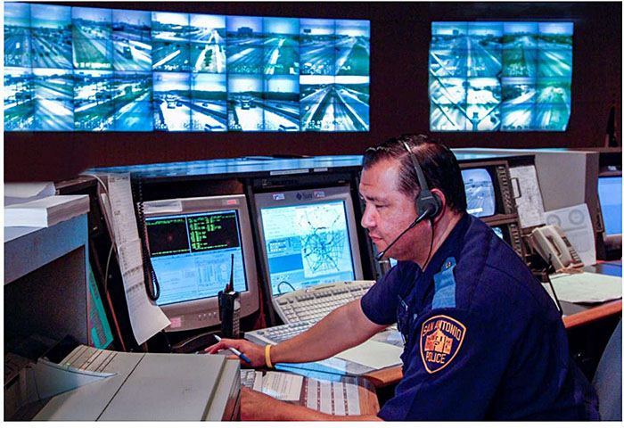 Photo of Transportation Management Center desk with TMC operator in front of various computers and multiple screens depicting roadway images from monitors in the transportation system.