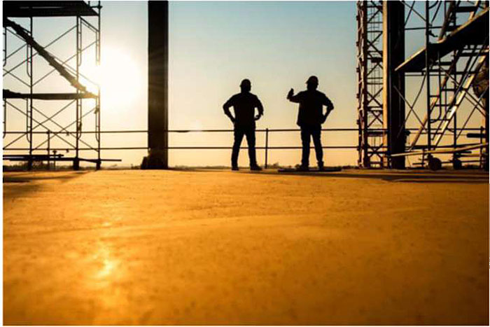 Stock photo of an outdoor industrial work site with silhouettes of two workers with hard hats talking to each other.